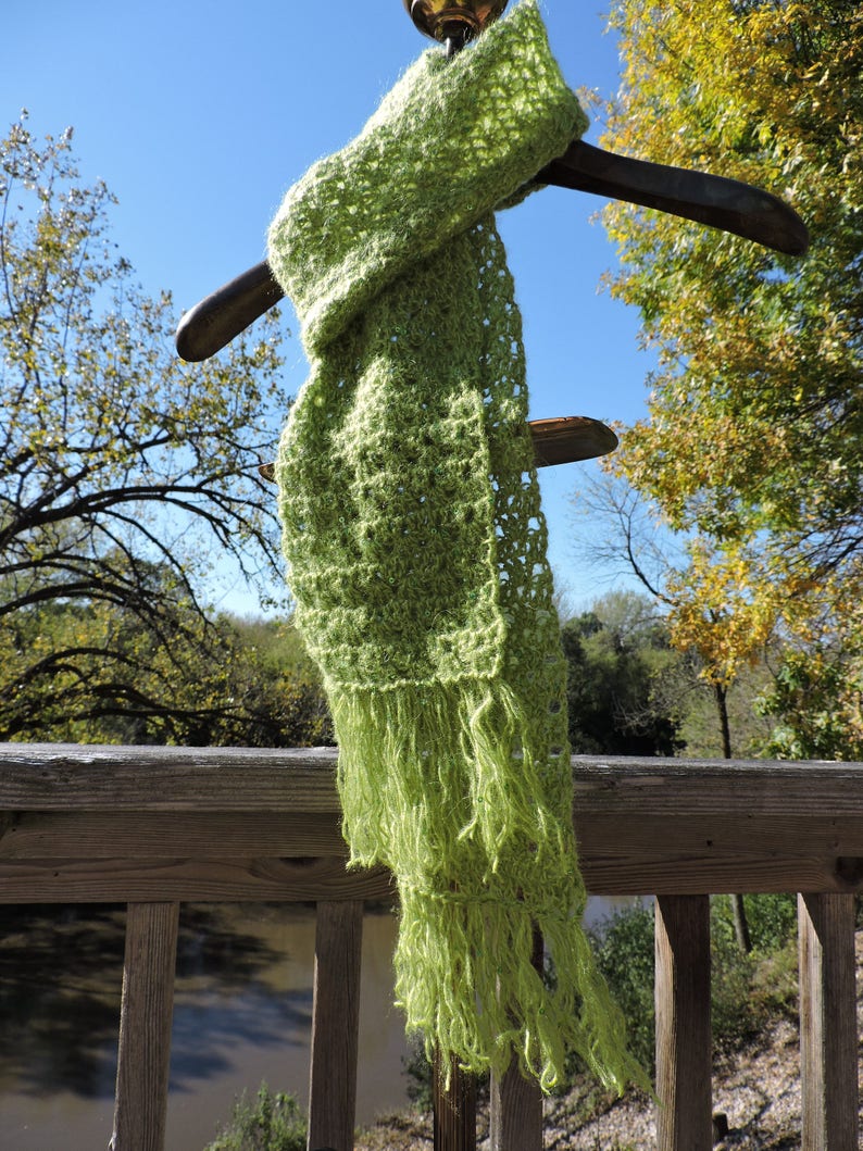 May include: A lime green crocheted scarf with fringe hanging from a wooden hanger. The scarf is displayed against a backdrop of a wooden railing and a body of water.