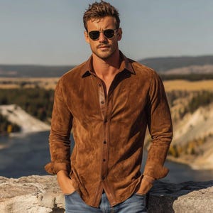 May include: A long-sleeved button-down shirt crafted from brown suede material, featuring a classic collar, a central placket with dark buttons, and a textured finish. The shirt is paired with blue denim jeans and dark-tinted aviator sunglasses.