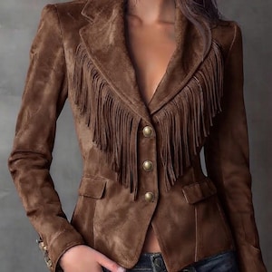 May include: A brown suede blazer featuring long fringe detailing along the lapels and chest area. The jacket includes three metallic buttons down the front, a notched collar, and two flap pockets at the waist, paired with dark blue denim jeans.