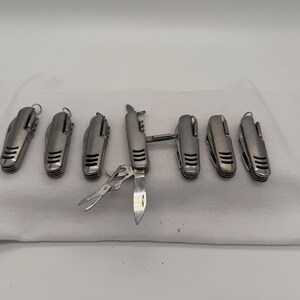 May include: Seven silver-toned multi-tool pocket knives arranged in a row. Six of the knives are in a closed position, while the central knife is partially open, revealing a small blade and a pair of scissors. Each handle features three horizontal slots.