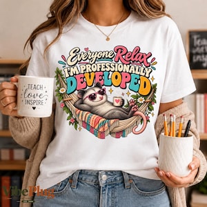 May include: A white t-shirt featuring a colourful graphic of an opossum wearing pink sunglasses, relaxing in a hammock while holding a mug. The text reads Everyone Relax I'M PROFESSIONALLY DEVELOPED. A white mug with the text TEACH love INSPIRE is held nearby.