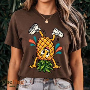 Könnte beinhalten: Ein dunkelbraunes Kurzarm-T-Shirt mit einer Grafik eines gelben Ananas-Charakters mit lächelndem Gesicht, der einen Handstand macht. Die Ananas trägt weiße Turnschuhe und ist von kleinen, bunten abstrakten Spritzern in Rot und Türkis umgeben.