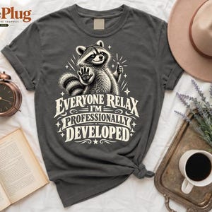 Puede incluir: Una camiseta gris carbón con un gráfico de un mapache con las patas levantadas y el texto "EVERYONE RELAX I'M PROFESSIONALLY DEVELOPED" en letras blancas. El diseño incluye pequeños detalles de estrellas que rodean la ilustración.