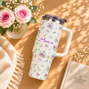 May include: A white stainless steel tumbler with a handle, featuring a repeating pattern of purple flowers, blue berries, and light blue butterflies. The name Maeve is printed in purple script on the front. The lid is clear plastic with a dark rim.