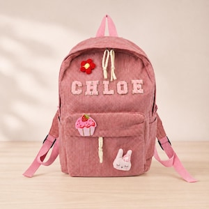 May include: A dusty rose corduroy backpack featuring the name CHLOE in light pink block letters. The bag includes a red flower patch, a pink cupcake patch, and a small white rabbit face patch. It has a top handle and two adjustable shoulder straps.