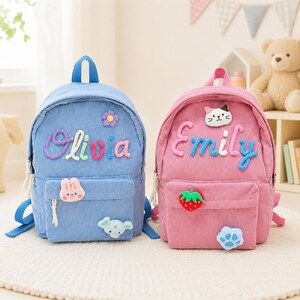 May include: Two corduroy backpacks, one blue and one pink. The blue backpack features the name Olivia in colorful letters, a purple flower, a pink rabbit, and a light blue elephant. The pink backpack features the name Emily, a cat, a strawberry, and a paw print.