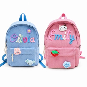 May include: Two corduroy backpacks, one light blue and one pink, each featuring a front pocket and a top handle. The blue backpack displays the name Olivia in colorful yarn letters with a flower, rabbit, and elephant patch. The pink backpack displays the name Emily in blue yarn letters with a cat, strawberry, and paw print patch.
