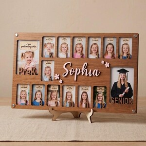 May include: A rectangular wooden school year photo frame with the name Sophia in pink script. It features thirteen photo slots labeled from PreK to senior year, with small pink flower accents and a stand for display on a flat surface.