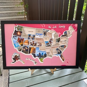 Puede incluir: Un mapa enmarcado de los Estados Unidos con fondo rosa y marco negro. El mapa presenta recortes de madera de los estados, algunos con fotografías personales. El texto "The Joh Family" está impreso en blanco en la parte superior derecha.