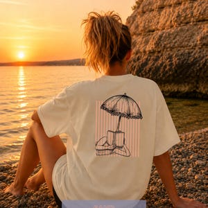 May include: A cream-colored t-shirt featuring a black line art graphic on the back. The illustration depicts a person lying down reading a book under a fringed beach umbrella, set against a background of vertical light pink stripes.