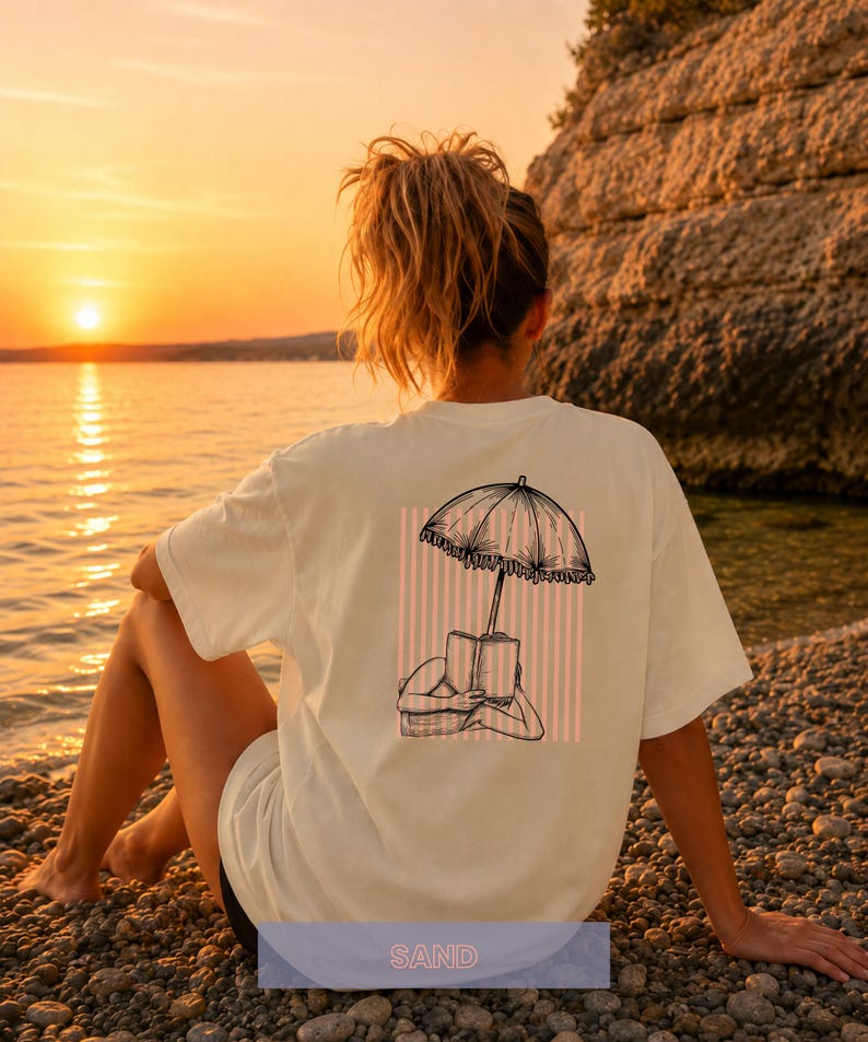 May include: A cream-colored t-shirt featuring a black line art graphic on the back. The illustration depicts a person lying down reading a book under a fringed beach umbrella, set against a background of vertical light pink stripes.
