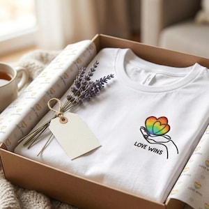 Könnte beinhalten: Ein weißes T-Shirt mit einer Stickerei, die eine Hand zeigt, welche ein regenbogenfarbenes Herz hält, darunter der Text LOVE WINS. Das T-Shirt liegt in einem Karton mit gemustertem Seidenpapier und einem Zweig getrocknetem Lavendel.