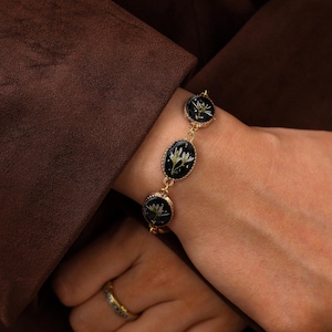 May include: A gold-toned chain bracelet featuring three oval-shaped charms with black resin centers, each containing a small white pressed flower. The bracelet is secured with a delicate gold metal link chain and has a polished, reflective finish.
