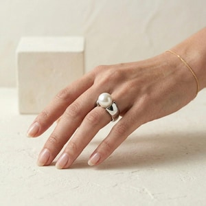 May include: A silver ring featuring a large, round, white pearl set in a wide, polished metal band. The ring is worn on a finger, and a thin gold chain bracelet is visible on the wrist. The design is characterised by its smooth, reflective surface.