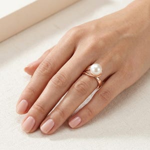 Peut inclure: Une bague en or rose ornée d'une perle blanche ronde unique, montée sur un anneau de style bypass. Le métal présente une finition lisse et polie, tandis que la perle est placée au centre, offrant un design classique à la surface lustrée.