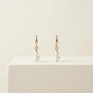 May include: A pair of gold drop earrings, each featuring two small, round white pearls stacked vertically. The pearls are suspended from a thin gold hook wire, creating a simple, elegant design that hangs downward with a polished finish.