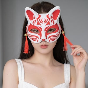 Op de afbeelding: Een wit vossenmasker met opvallende rode grafische patronen rond de ogen en oren. Het masker is voorzien van twee goudkleurige kralen bevestigd aan lange, felrode kwastjes die aan beide zijden hangen en het bovenste deel van het gezicht bedekken.