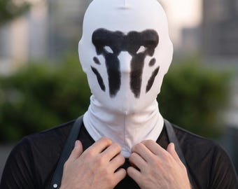 May include: A white full-head mask featuring a black inkblot pattern across the face area. The mask is made of a smooth, form-fitting fabric that covers the head and neck, displaying a symmetrical, abstract design reminiscent of a Rorschach test.