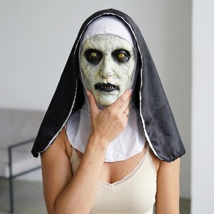 May include: A full-head nun mask featuring a pale, cracked skin texture, deep-set yellow eyes, and dark, sunken features. The mask is attached to a traditional black and white habit headpiece that covers the head and shoulders in a classic style.