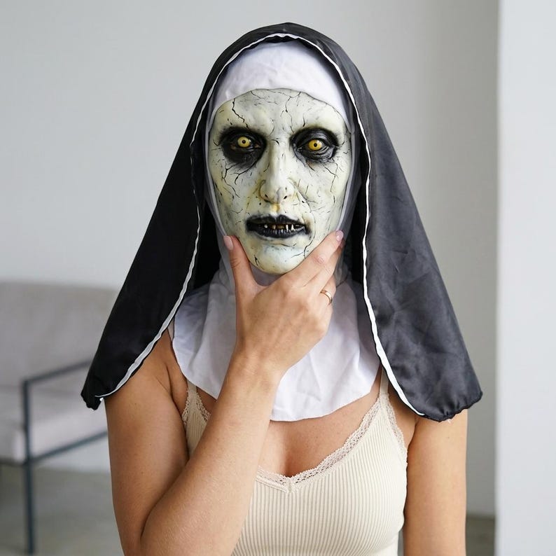 May include: A full-head nun mask featuring a pale, cracked skin texture, deep-set yellow eyes, and dark, sunken features. The mask is attached to a traditional black and white habit headpiece that covers the head and shoulders in a classic style.