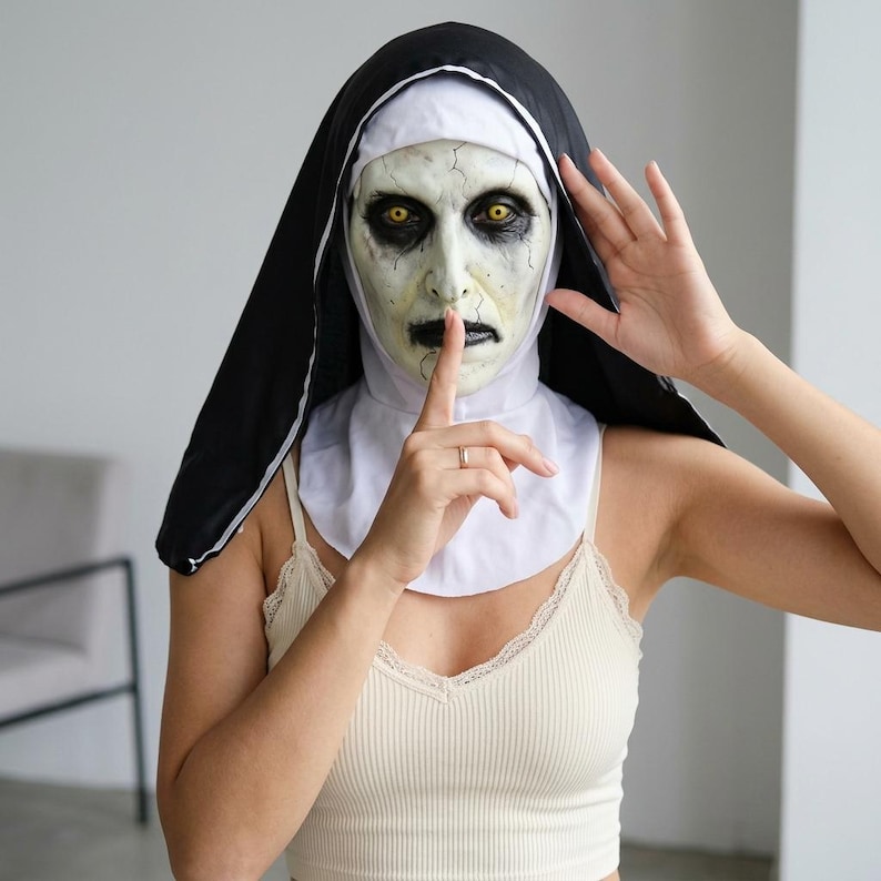 May include: A full-head nun mask featuring a pale, cracked face with dark sunken eyes, yellow irises, and black lips. The mask includes a traditional black and white habit headpiece that covers the head and neck, creating a haunting appearance.