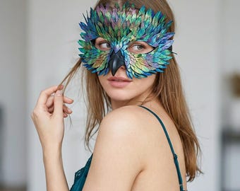 May include: A masquerade mask shaped like an owl face, featuring iridescent green, blue, and purple layered feather-like scales. The mask includes a prominent, smooth black beak at the center and covers the eye area with a textured, metallic finish.