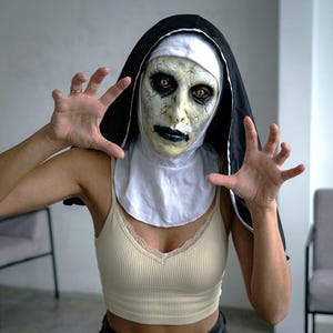May include: A nun mask featuring a pale, cracked face with dark sunken eyes and black lips, paired with a traditional black and white habit. The mask covers the head and neck, worn over a beige ribbed tank top with lace trim along the neckline.