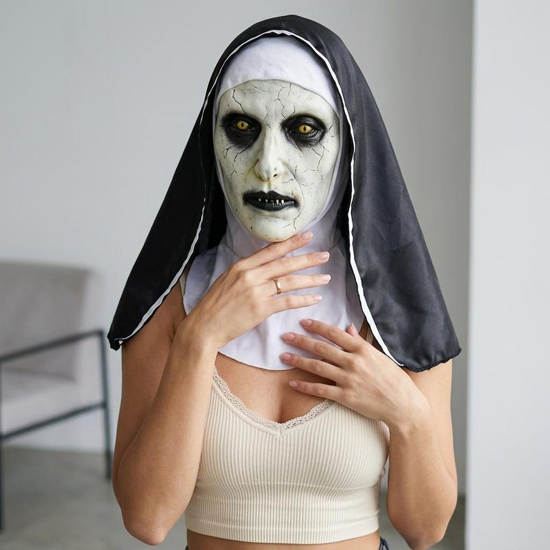 May include: A full-head nun mask featuring a pale, cracked face with deep-set yellow eyes, dark eye makeup, and black lips. The mask includes an attached black and white habit headpiece that covers the head and neck, creating a realistic appearance.