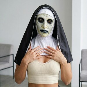 May include: A full-head nun mask featuring a pale, cracked, and weathered face with dark, sunken eyes and black lips. The mask includes an attached black and white habit headpiece that drapes over the shoulders, creating a realistic horror costume.
