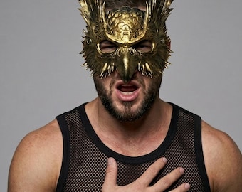 May include: A metallic gold-colored mask shaped like an eagle or bird of prey with intricate feather detailing and a sharp beak. The mask covers the upper portion of the face, featuring cutouts for the eyes and a textured, sculpted surface finish.
