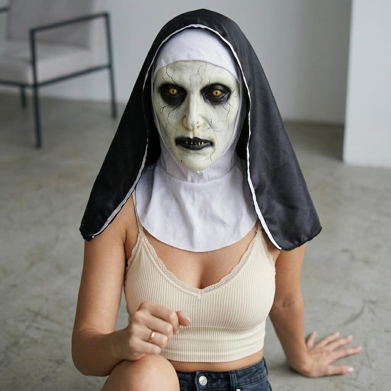 May include: A full-head nun mask featuring a pale, cracked face with dark sunken eyes, yellow irises, and black lips. The mask includes an attached black and white habit headpiece that covers the head and neck, creating a stark, high-contrast look.