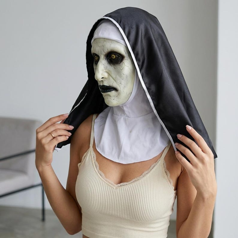 May include: A full-head nun mask featuring a pale, cracked skin texture, deep-set yellow eyes, and dark lips. The mask is attached to a black fabric habit with a white wimple that covers the head and neck, creating a stark, high-contrast appearance.