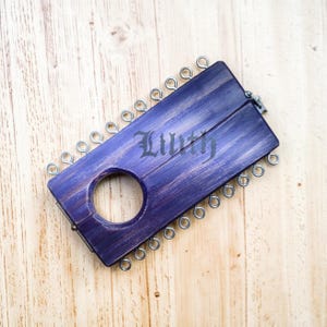 May include: A purple wooden object with a circular hole and metal rings. The object has the word "Lilith" printed on it. The wood grain is visible, and the object is set against a light-colored wooden surface.