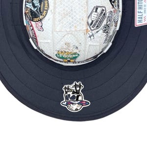 May include: A white hard hat with a wide dark navy blue brim and various stickers, including one that says YOUNGSTOWN. A graphic of a skull wearing a hat is centred on the brim. A tag on the side reads HALF HITCH SINCE 22.