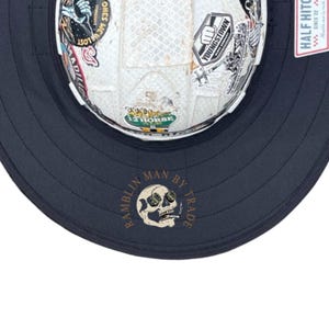 May include: A white hard hat covered in various colourful stickers, featuring a wide black brim. The brim displays a circular logo with a skull graphic and the text "RAMBLIN MAN BY TRADE" in gold lettering. A small rectangular tag is attached to the side.