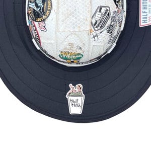 May include: A white hard hat with a wide dark navy brim, covered in various colourful stickers. A small sticker on the front brim depicts a person in a bathtub with the text Half Hitch. A tag on the side reads Half Hitch Since 22.