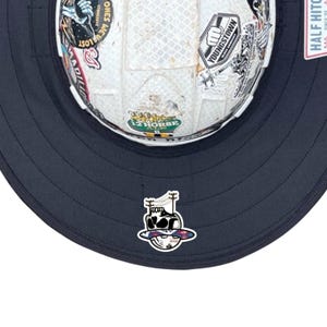 May include: A white hard hat featuring a wide dark navy brim and various stickers, including one with a skull design on the front brim. The hard hat displays multiple decals such as Youngstown Glove Company and 12 Horse Ale logos on its surface.