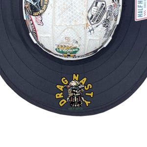 May include: A dark navy blue wide-brimmed hat featuring a white hard hat covered in various colourful stickers at the centre. The brim displays a graphic of a skull wearing a hard hat with the yellow text DRAG NASTY and green text HALF HITCH.