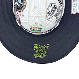 May include: A white hard hat with a textured surface covered in various colourful stickers, featuring a wide black brim. The underside of the brim displays the green text "This ain't daddy's money" in a stylised script font, with a small tag attached.