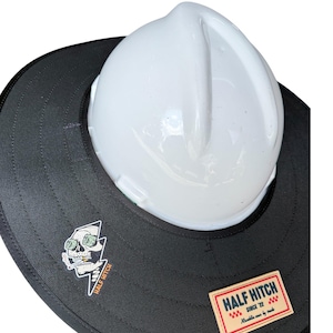 May include: A white hard hat with a wide black brim attachment. The brim features a graphic of a skull with money eyes smoking a cigarette and a rectangular tan patch with the text HALF HITCH SINCE '22 and the phrase Ramblin men by trade.