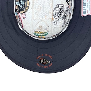 May include: A white hard hat with a textured surface covered in various colourful stickers, including one reading YOUNGSTOWN. The hat features a wide, dark navy brim with a graphic of a skeleton hand holding a rose and the text DEATH BEFORE SHITTY PER DIEM.