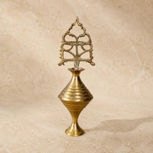 May include: A metallic gold-toned vessel with a conical body featuring horizontal ridges, a narrow neck, and a flared base. The vessel is topped with an intricate, openwork metal finial that has a pointed, triangular silhouette and ornate cutouts.
