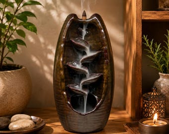 May include: A dark brown ceramic backflow incense burner with a tall, oval shape and a ribbed base. The burner features a series of cascading tiers that guide white incense smoke downward in a flowing stream from a lit cone at the top.