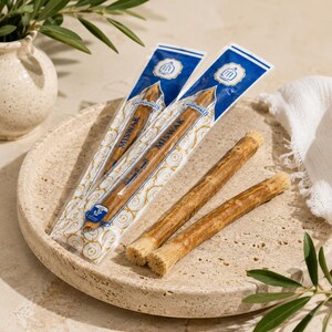 May include: Two miswak sticks in clear plastic packaging with blue and gold accents, featuring the text MISWAK, alongside two unwrapped miswak sticks with fibrous, brush-like ends. The sticks are light brown with a natural, woody texture.