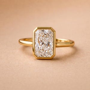 May include: A gold ring featuring a large, rectangular radiant-cut clear gemstone set in a smooth, polished bezel setting. The band is thin, rounded, and crafted from a solid yellow gold metal, providing a clean and simple frame for the center stone.