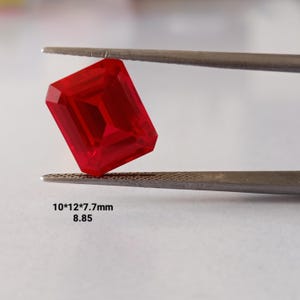 May include: A vibrant red, emerald-cut gemstone held by metal tweezers. The stone features a faceted surface with sharp, clean edges. Printed text below the stone indicates dimensions of 10 by 12 by 7.7mm and a weight value of 8.85.
