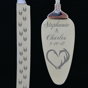 May include: A silver cake knife and server set with engraved designs. The server has a heart with antlers and the text "Stephanie & Charles 8-10-17". The knife has a pattern of deer tracks.