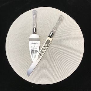 May include: A set of two silver cake knives with clear plastic handles. The larger knife has the text "Jennifer & Matthew 6-29-2019" and an American flag design engraved on the blade.