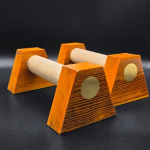 Könnte beinhalten: Ein Paar Liegestützgriffe aus Holz mit hellen, zylindrischen Griffen, die mit zwei trapezförmigen Sockeln verbunden sind, welche eine dunkelorangefarbene Holzmaserung und eine kreisförmige, goldfarbene Metalleinlage auf der Vorderseite aufweisen.