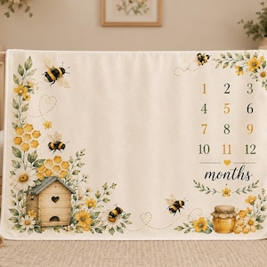 May include: Cream-colored baby milestone blanket with a bee theme. Features illustrations of bees, honeycombs, flowers, and a wooden beehive. Includes numbers 1-12 for monthly photos. A jar of honey is also shown.